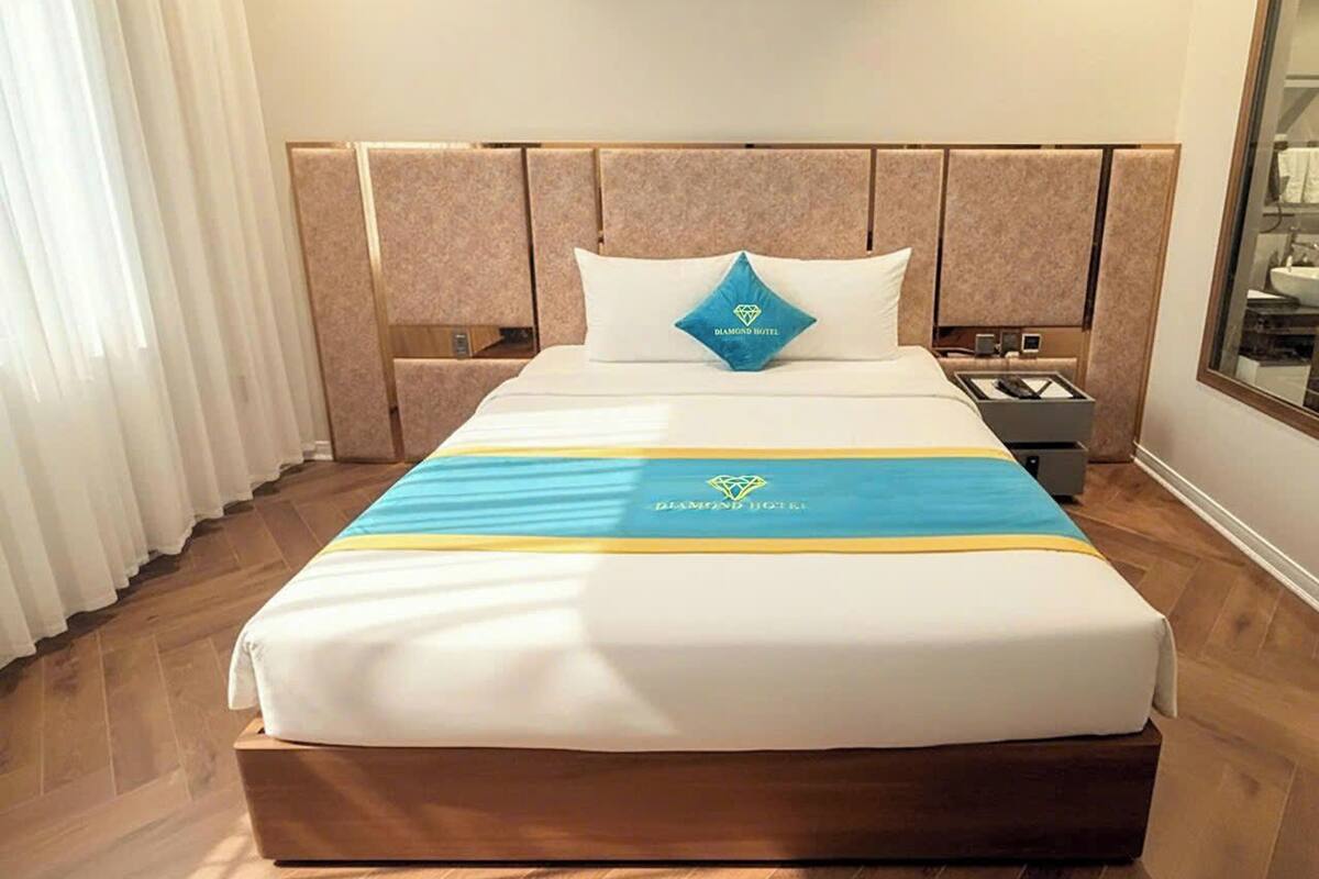 A hotel room with a large bed, a headboard with a diamond-shaped logo, a bedside table, and a mirror.