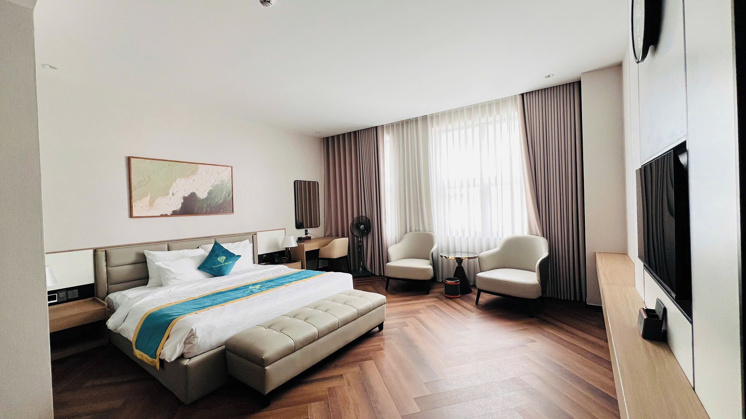 Una habitación de hotel con una cama, dos sillones, un televisor y un ventanal con cortinas.