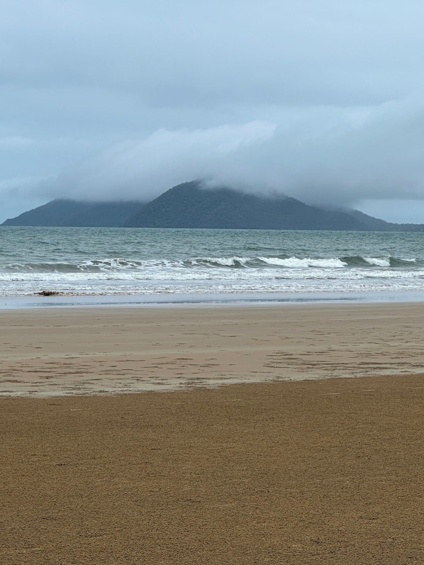 Una playa con olas, una orilla de arena y una montaña parcialmente cubierta de nubes al fondo.
