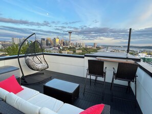 Eine Dachterrasse mit einer Couch, Stühlen und einem Hängesessel mit Blick auf eine Stadtlandschaft und einem markanten Turm.