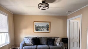 Un salon avec un canapé gris, une table basse noire, un tapis à motifs, un ventilateur de plafond et un tableau représentant un paysage, encadré et accroché au mur.