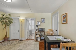 A room with a dining table, chairs, a microwave, a bookshelf, a potted plant, and a door leading to another room.