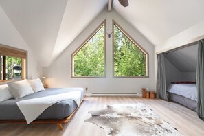 Ein Schlafzimmer mit schrägem Dach, einem Bett mit grauer Matratze, einer weißen Tagesdecke, Holzboden, einem großen Fenster mit Blick auf Bäume und einem Fellteppich.