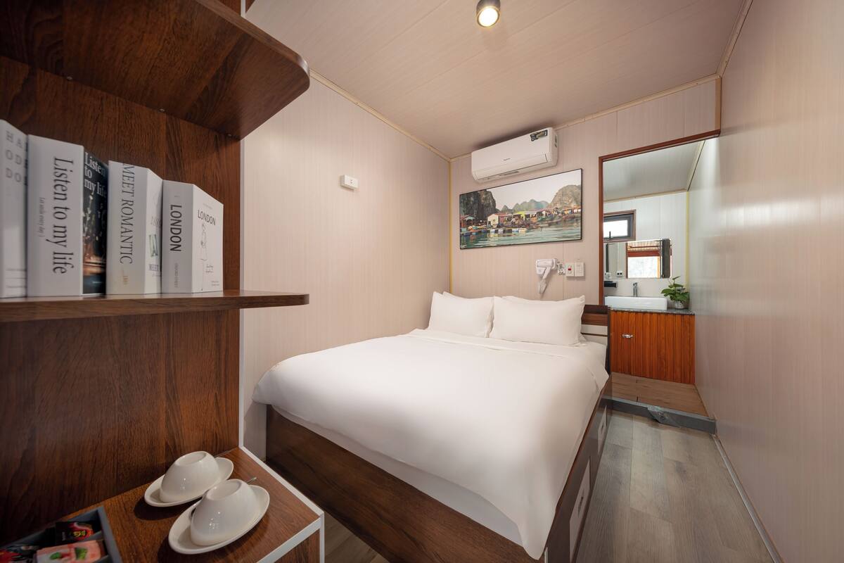 A hotel room with a bed, a wooden shelf with books, a small table with a pair of shoes, a mirror, and a wall-mounted air conditioner.