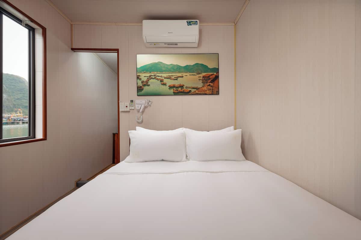 A hotel room with a bed, a mounted air conditioner, a painting of a landscape, and a telephone.