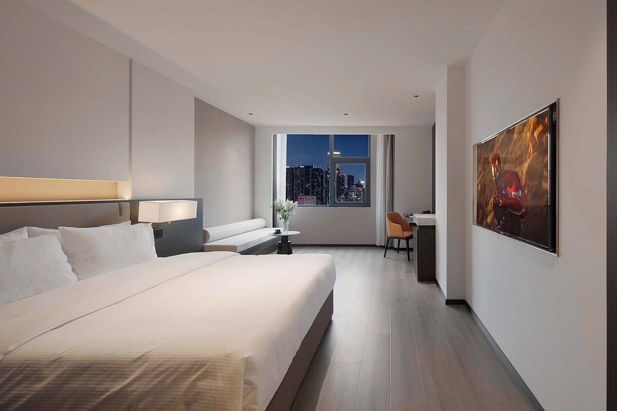 A hotel room with a large bed, a desk, a chair, a window with a city view, and a painting on the wall.