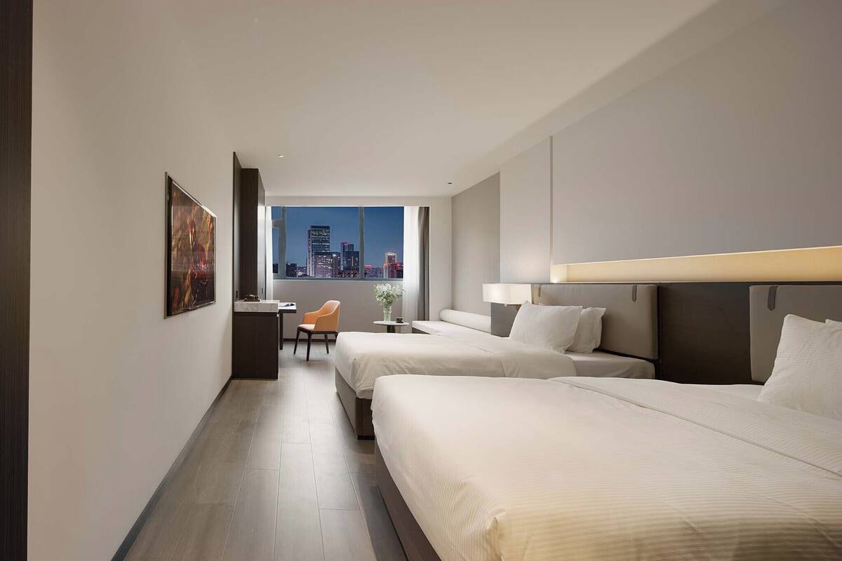 A hotel room with two beds, a desk, a chair, and a large window with a city view.