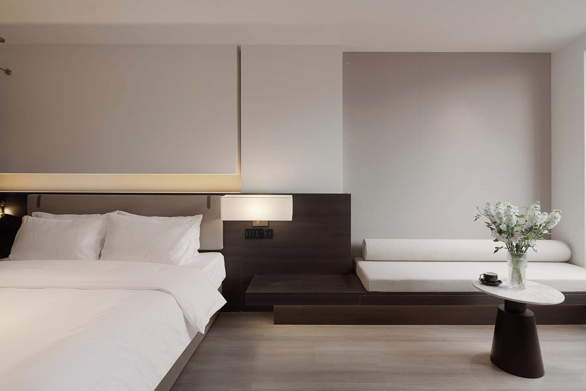 A modern hotel room with a bed, bedside table, lamp, and a small seating area with a vase of flowers.