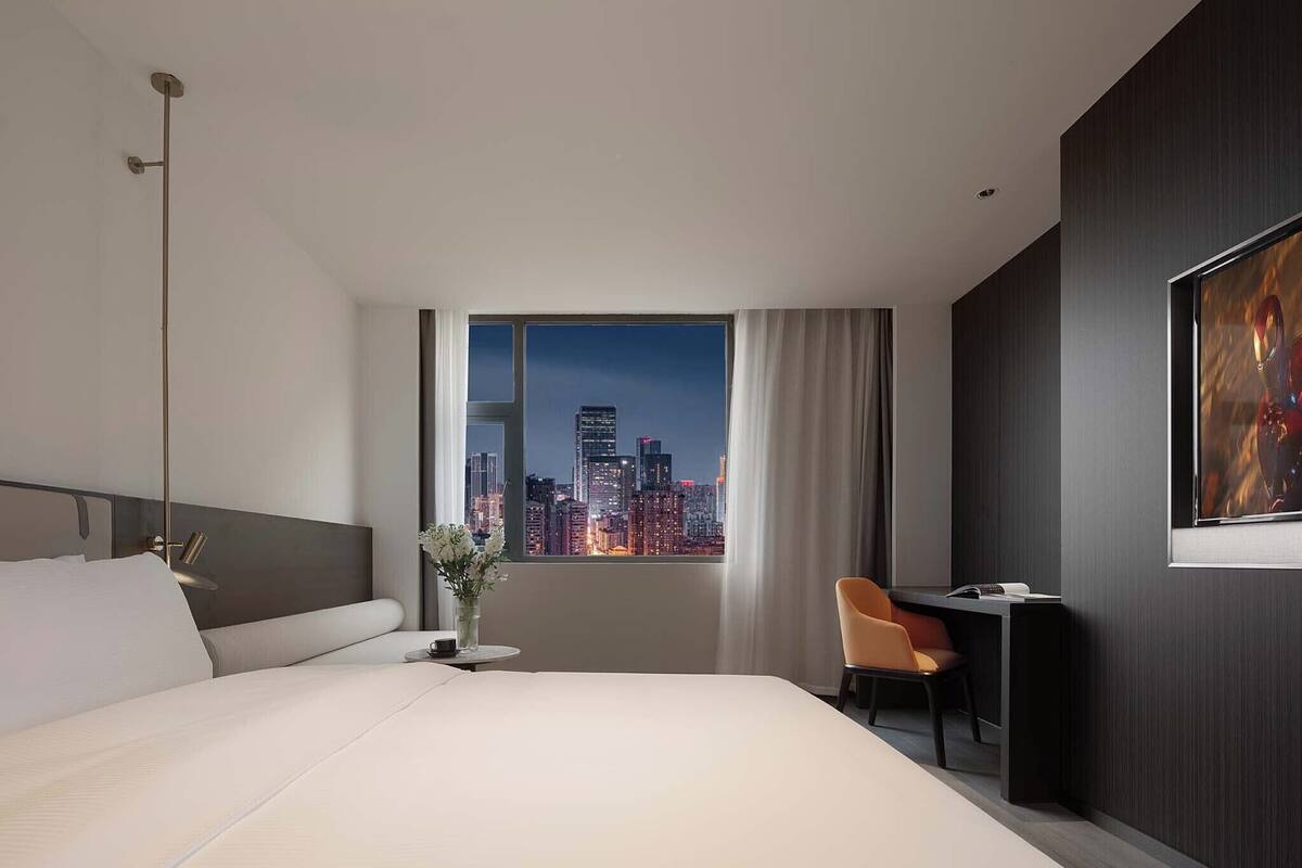 A modern hotel room with a large bed, a desk, a chair, a window with a cityscape view, and a painting on the wall.
