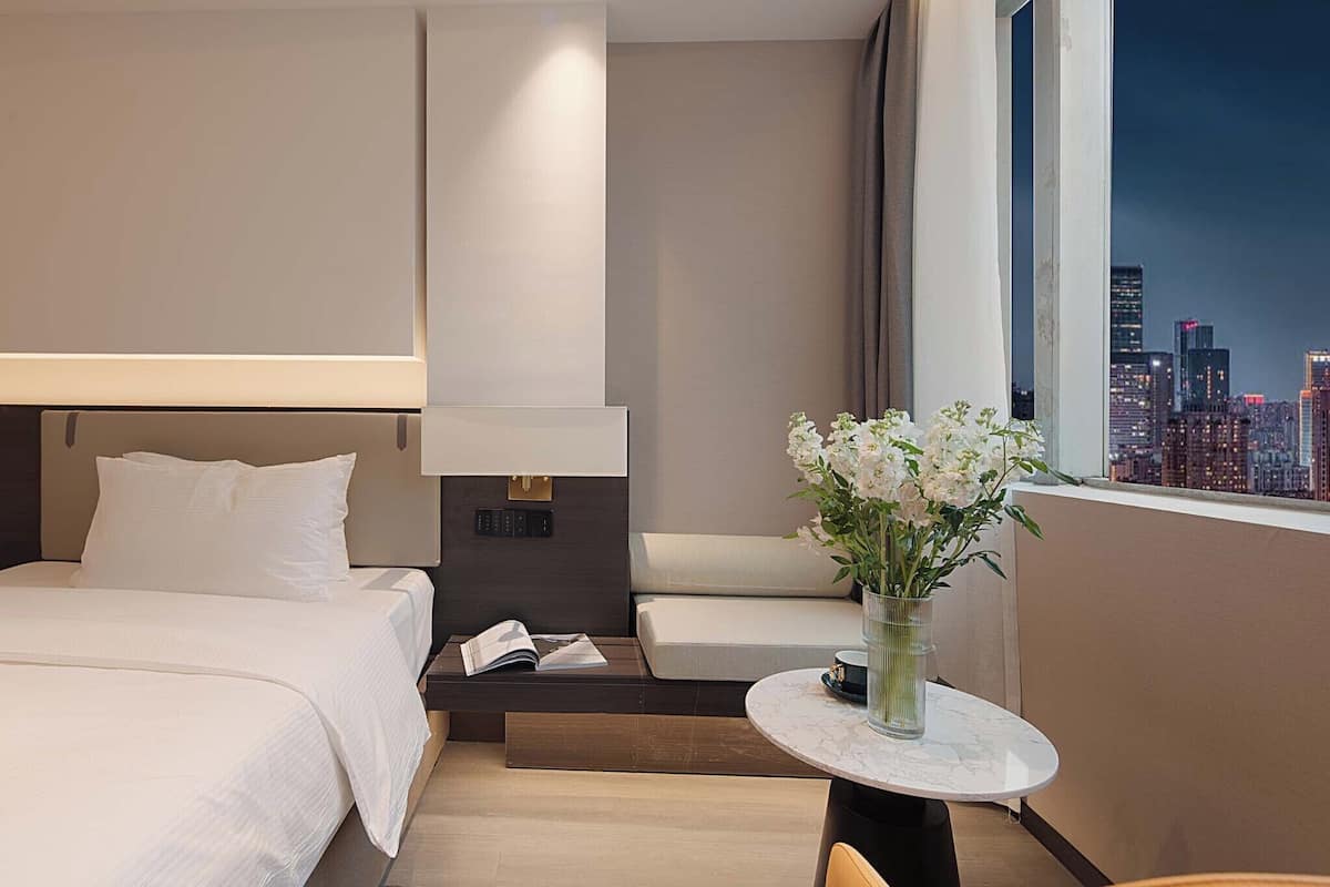 A modern hotel room with a bed, bedside table, a small round table with a vase of flowers, and a view of a cityscape.