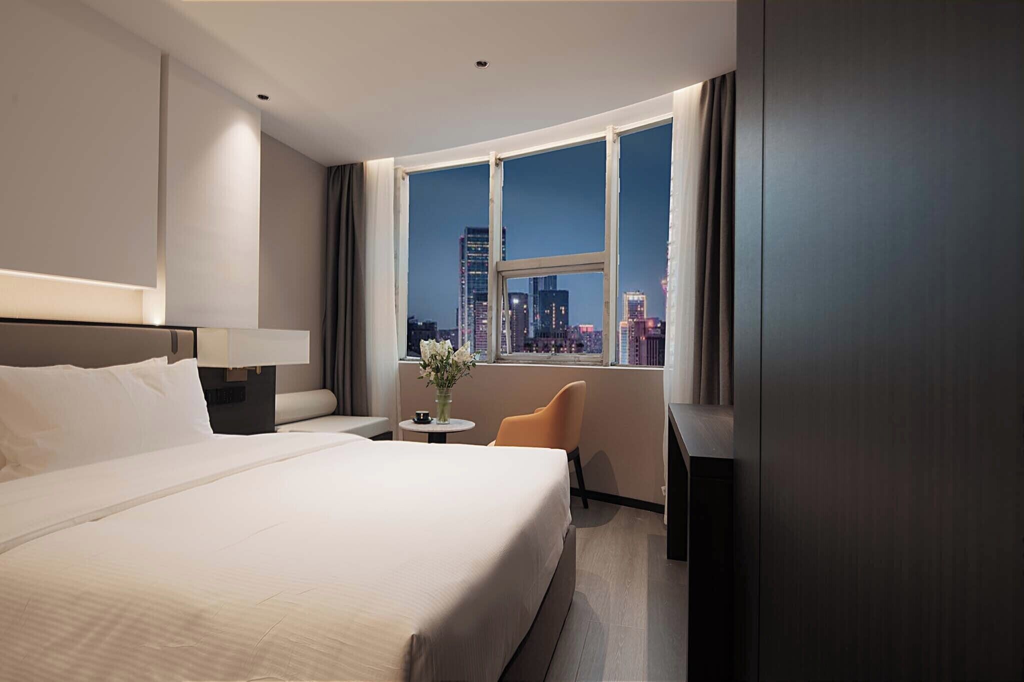 A hotel room with a large bed, a small table with a vase, a chair, and a view of a cityscape through a large window.
