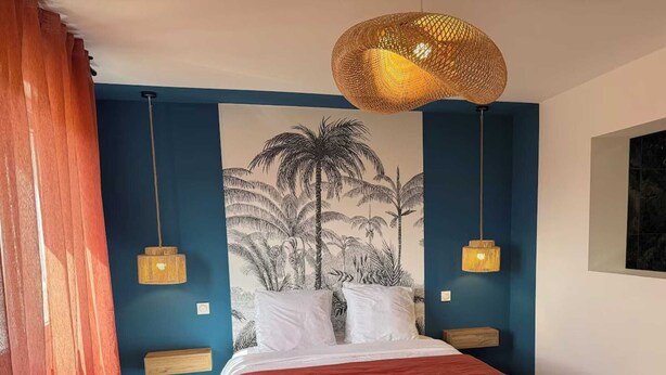Une chambre à coucher comprenant un lit, deux tables de chevet, un luminaire suspendu, un banc et une fresque murale.