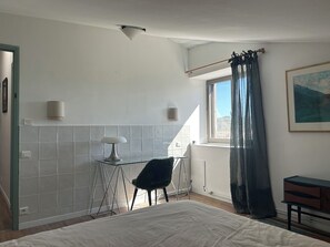 Ett sovrum med en säng, en stol, ett litet bord, ett fönster med gardiner och en tavla på väggen.