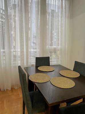 Ruokapöytä, jonka päällä on neljä punottua alustaa, ja pöydän ympärillä tuoleja. Huoneessa on läpikuultavat verhot, ja ikkunasta avautuu näkymä ulkona oleviin rakennuksiin.