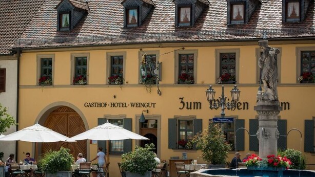 A building with a yellow facade, a large arched entrance, outdoor seating with umbrellas, and a statue in the foreground. - Gasthof zum weißen Lamm (Sommerach)