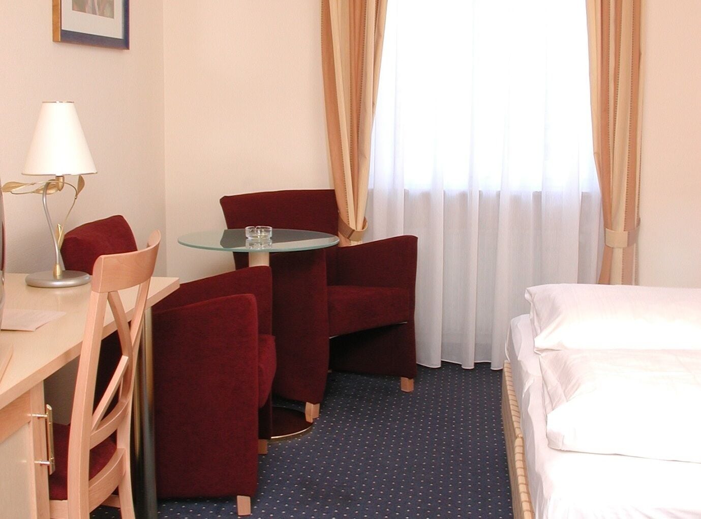 Habitación de hotel con cama, escritorio con lámpara, silla, mesita y ventana con cortinas.