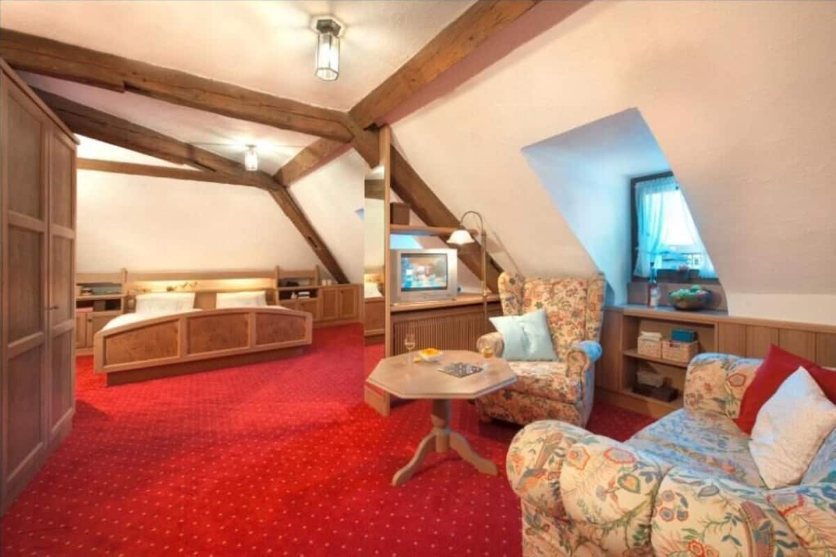 A cozy attic room with a sloped ceiling, a bed, a sofa, a small table, and a television.