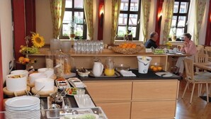A breakfast buffet with various food items and beverages, a coffee station, and a seating area with two people dining. - Gasthof zum weißen Lamm (Sommerach)