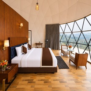 A modern hotel room with a large bed, wooden panel walls, a view through a large window, and a small seating area. - LushGreen Kodai by Arcadia (Kodaikanal)