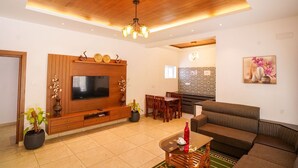 A modern living room with a wooden entertainment unit, a flat-screen TV, a sofa, a dining area, and a kitchen in the background.