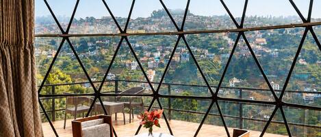 Un dormitorio con una cama grande, una mesita con un florero, dos sillas y vista a un paisaje urbano a través de una ventana de diseño geométrico.