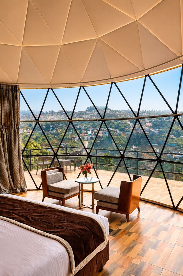 A bedroom with a large bed, a small table with a vase, two chairs, and a view of a cityscape through a geometric-patterned window. - LushGreen Kodai by Arcadia (Kodaikanal)
