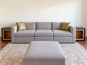 A grey sofa with two cushions, a matching ottoman, and two side tables with wooden frames. - 2BR Traveler / Domain Louis Vuitton Building (Austin)