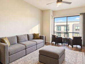 A modern living room with a grey sofa, ottoman, and wicker chairs. A large window offers a view of the building's exterior. - 2BR Traveler / Domain Louis Vuitton Building (Austin)