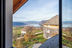 A view from a window overlooking a rural landscape with a wooden house, a body of water, and distant mountains. - Villa overlooking Lake Annecy (Saint-Eustache)