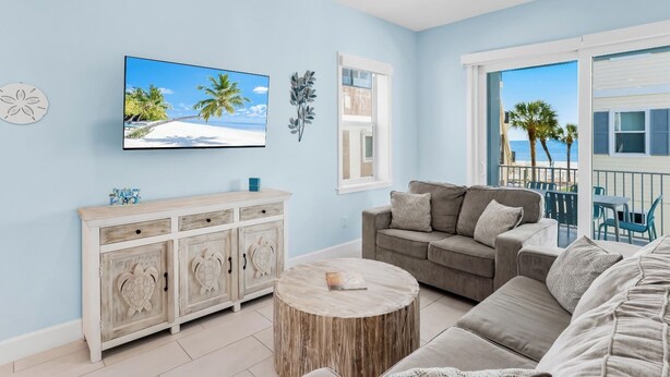 Uma sala de estar com sofá, mesa de centro redonda, aparador com detalhes decorativos, televisão e janela com vista para a praia.