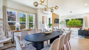 A modern dining area with a large oval table, multiple chairs, a chandelier, and large windows offering a view of greenery outside. - AC / New Remodel / Chef's Kitchen / BBQ / Cap Hill (Seattle)