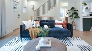 A modern living room with a dark blue sofa, a round coffee table with a book and a vase, a staircase, and a kitchen area visible in the background. - AC / New Remodel / Chef's Kitchen / BBQ / Cap Hill (Seattle)