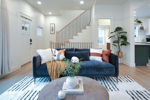 A modern living room with a dark blue sofa, a round coffee table with a book and a vase, a staircase, and a kitchen area visible in the background. - AC / New Remodel / Chef's Kitchen / BBQ / Cap Hill (Seattle)