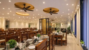 A dining area with multiple tables and chairs, ceiling fans, recessed lighting, and a view of a hallway with a blue-striped wall. - Hotel Paragon Suites (New Delhi)
