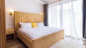 Ein modernes Schlafzimmer mit einem hölzernen Kopfteil, einem Einzelbett, einem Nachttisch und einem großen Fenster mit Vorhängen.