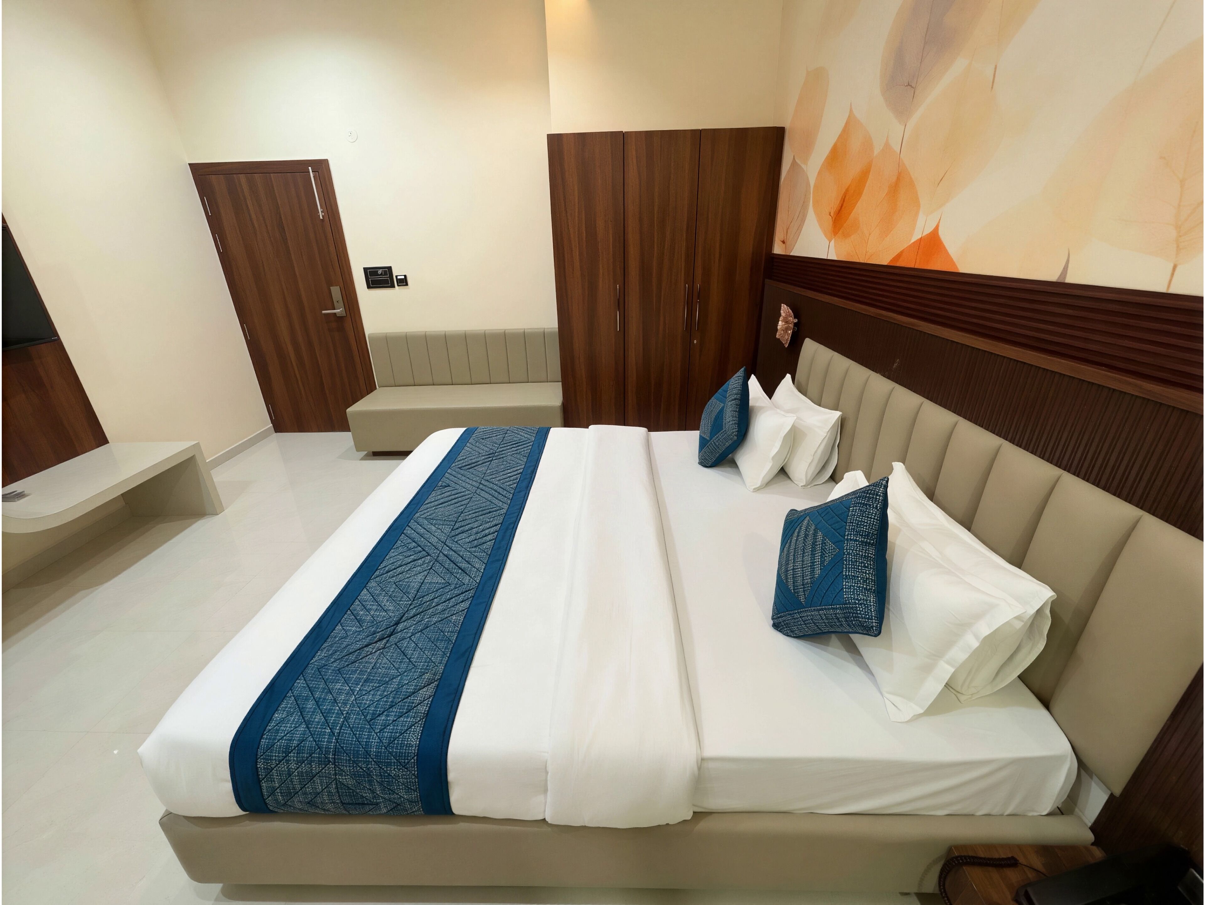 A neatly made bed with a blue and white bedspread, a wooden wardrobe, a bench, and a wall with a leaf pattern.