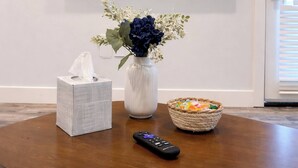Een televisie aan een muur bevestigd, toont 'Serenity Pines', een doos tissues, een vaas met bloemen, een mand met snacks en een afstandsbediening op een houten tafel.