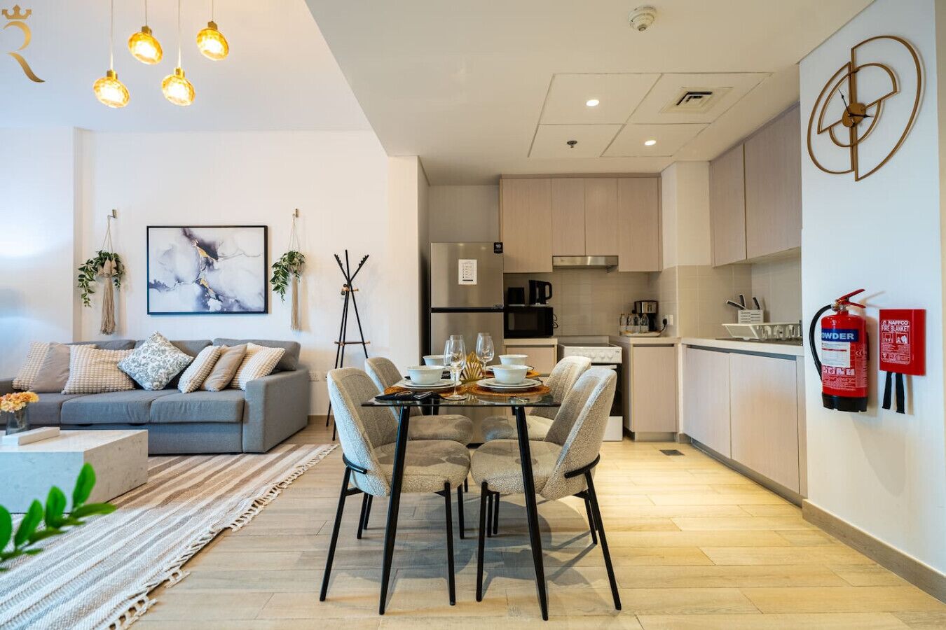 A modern living room with a grey sofa, a dining table with chairs, a kitchen area with cabinets, and a fire extinguisher mounted on the wall.