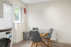 Een eethoek met een ronde glazen tafel en vier grijze stoelen, een wasmachine en een radiator.