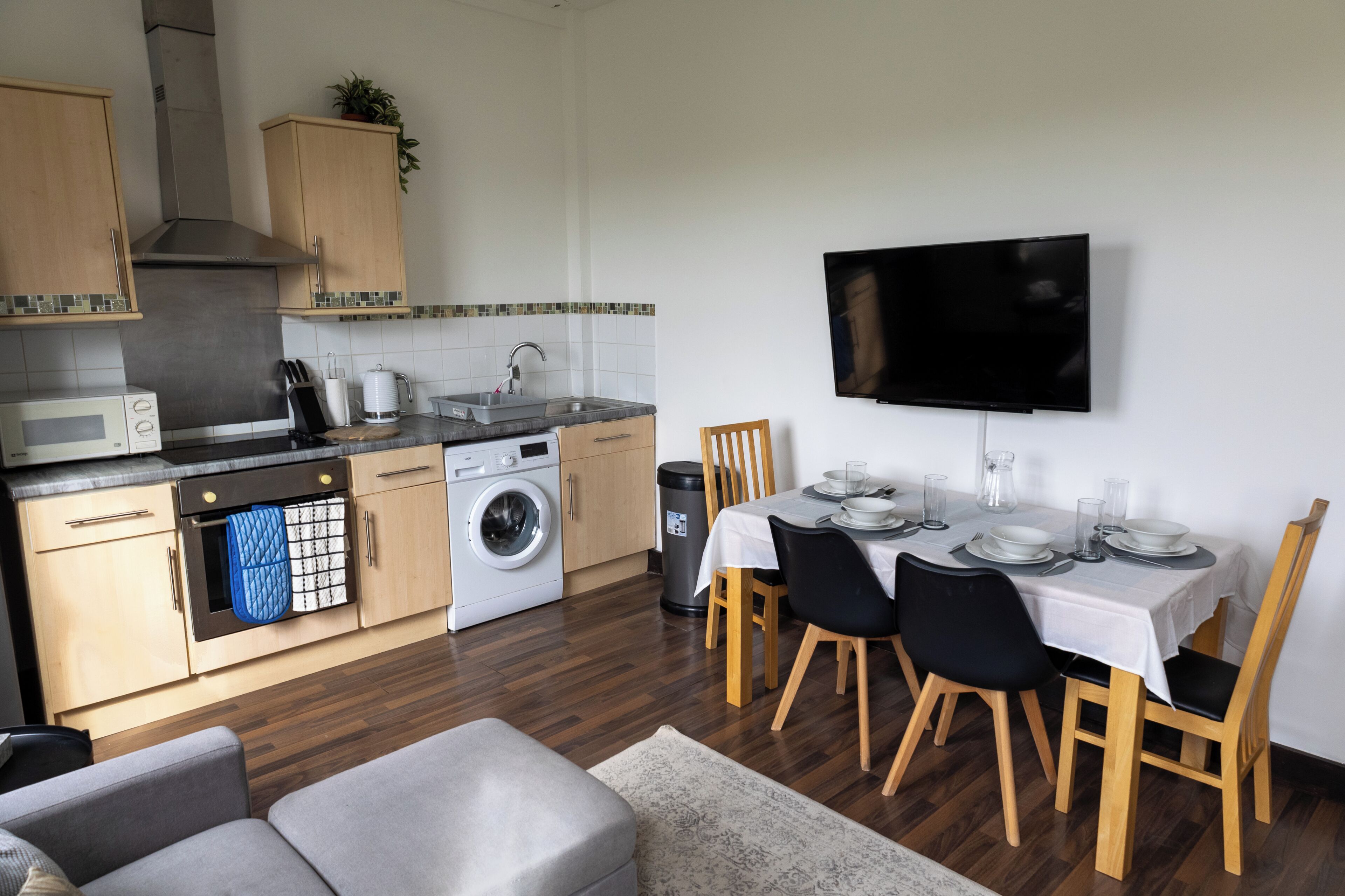 A compact kitchen with a washing machine, a sink, and a dining area with a TV above the table.