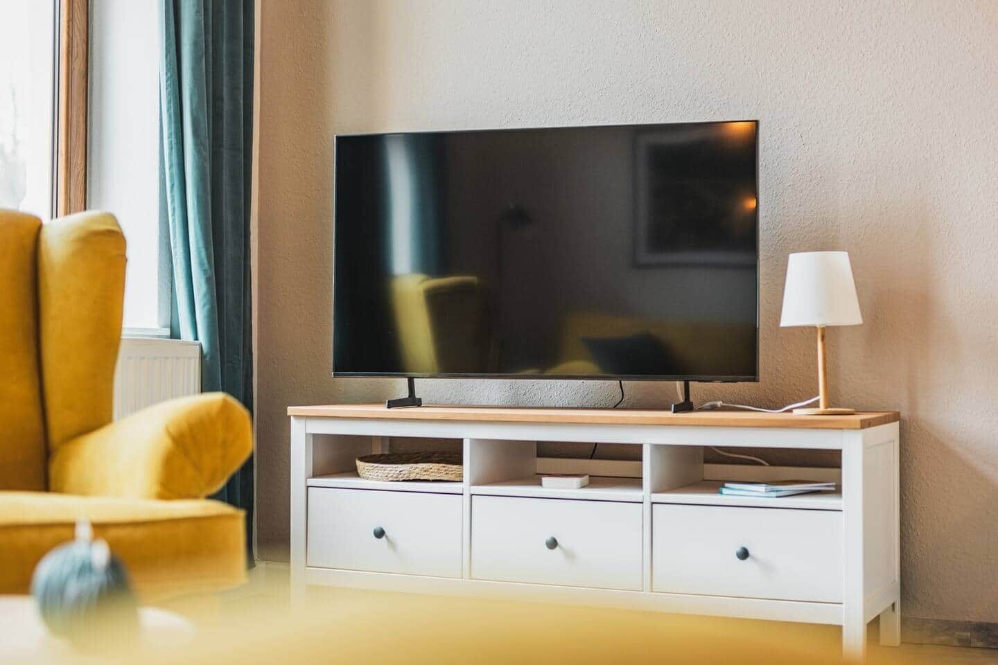 Ett modernt vardagsrum med en platt-TV på ett träställ, en gul fåtölj, ett vitt skåp med lådor och en lampa till höger.