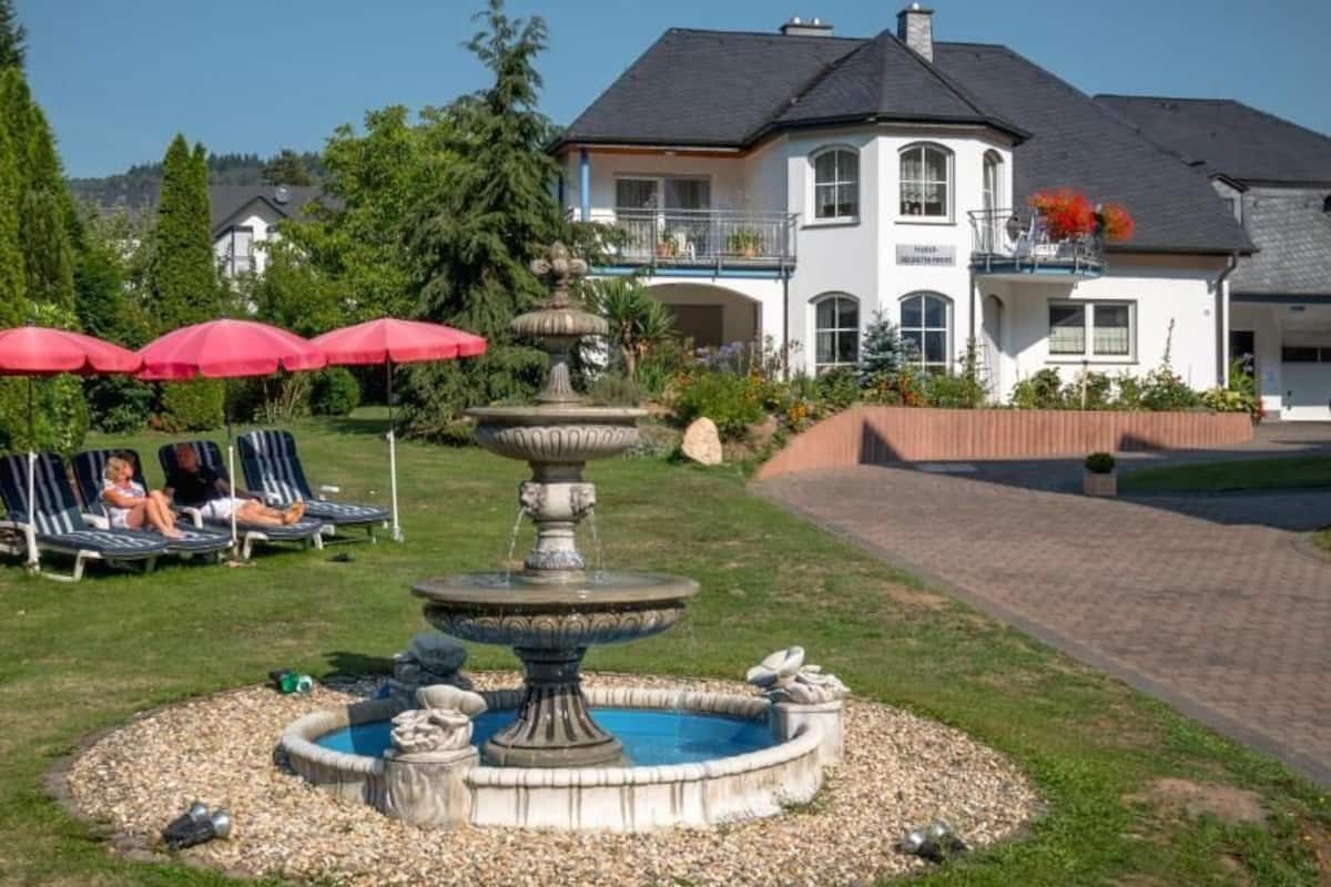 A fountain in a circular stone basin surrounded by a lawn, with red umbrellas and lounge chairs in the background.