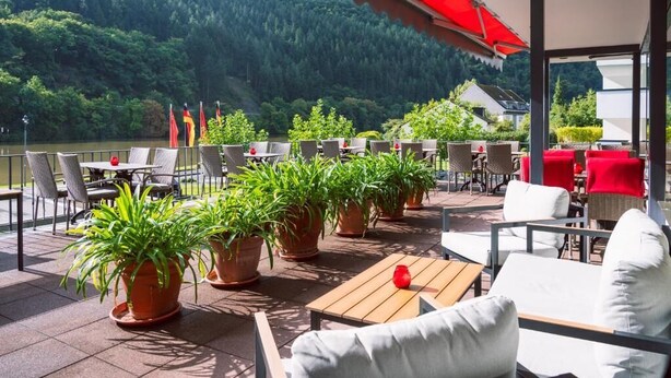 An outdoor patio with tables and chairs, potted plants, and a view of a lake and forest. - Hotel Bisenius (Traben-Trarbach)