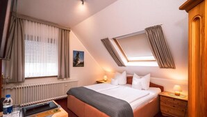 A hotel room with a bed, bedside tables, a desk with a chair, a radiator, a window with curtains, a skylight, and a wooden wardrobe. - Hotel Bisenius (Traben-Trarbach)