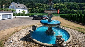A fountain with a tiered design, surrounded by pebbles, with a house and cars in the background. - Hotel Bisenius (Traben-Trarbach)