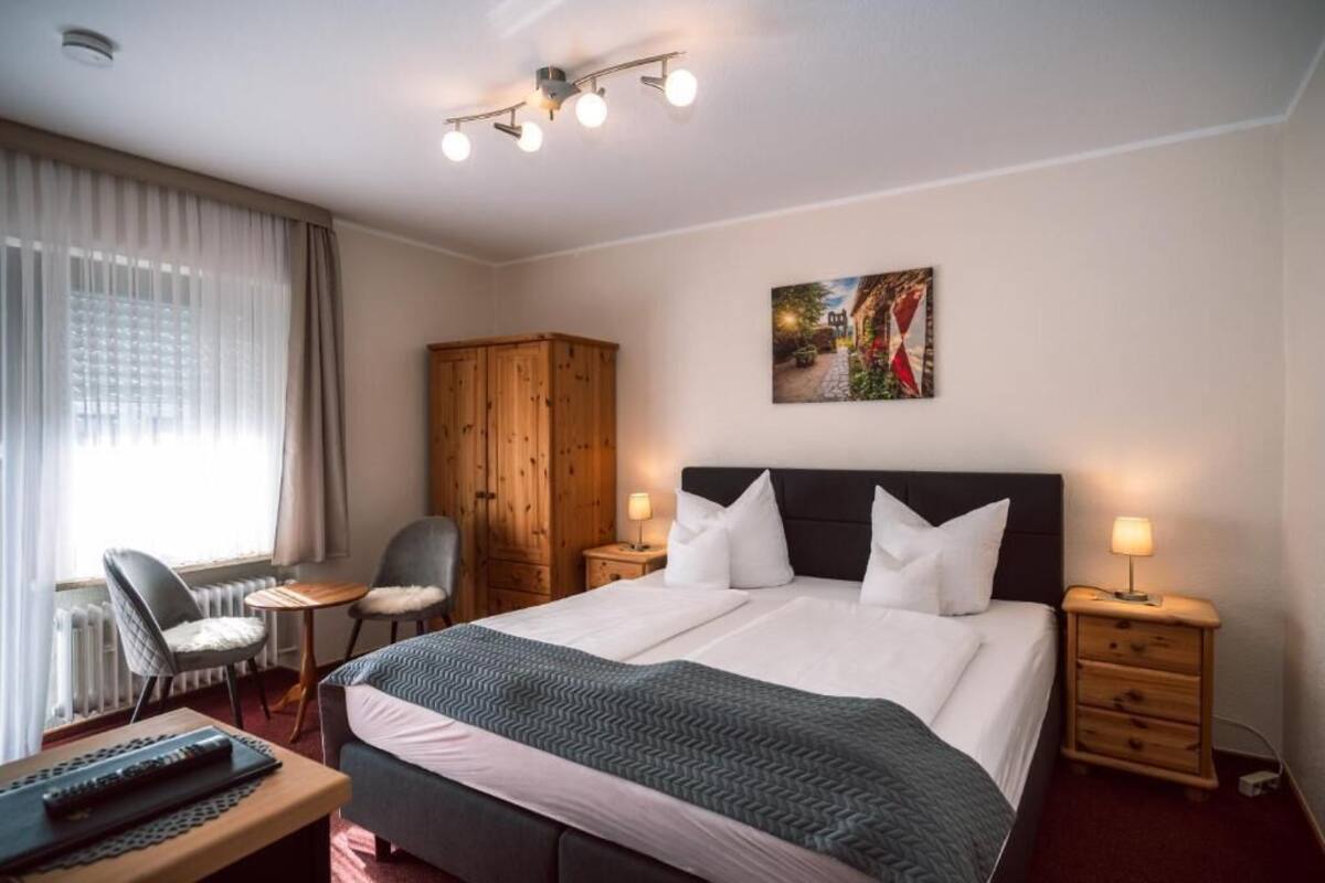 A hotel room with a bed, two bedside tables, a chair, a desk, a wooden wardrobe, and a painting on the wall.