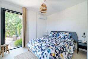 Un dormitorio con una cama grande, un ventilador de techo, un equipo de aire acondicionado, un taburete pequeño y vistas a la vegetación exterior.