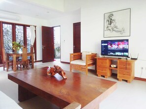 A spacious living area with a wooden dining table, a sofa, a TV, and a stone wall. - Malathi Private Suite Ubud - Green View Shared Pool (Ubud)