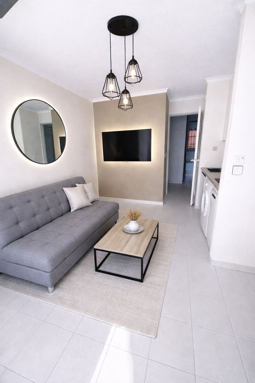 Un salon moderne avec un canapé gris, une table basse, un téléviseur à écran plat fixé au mur et un miroir rond.