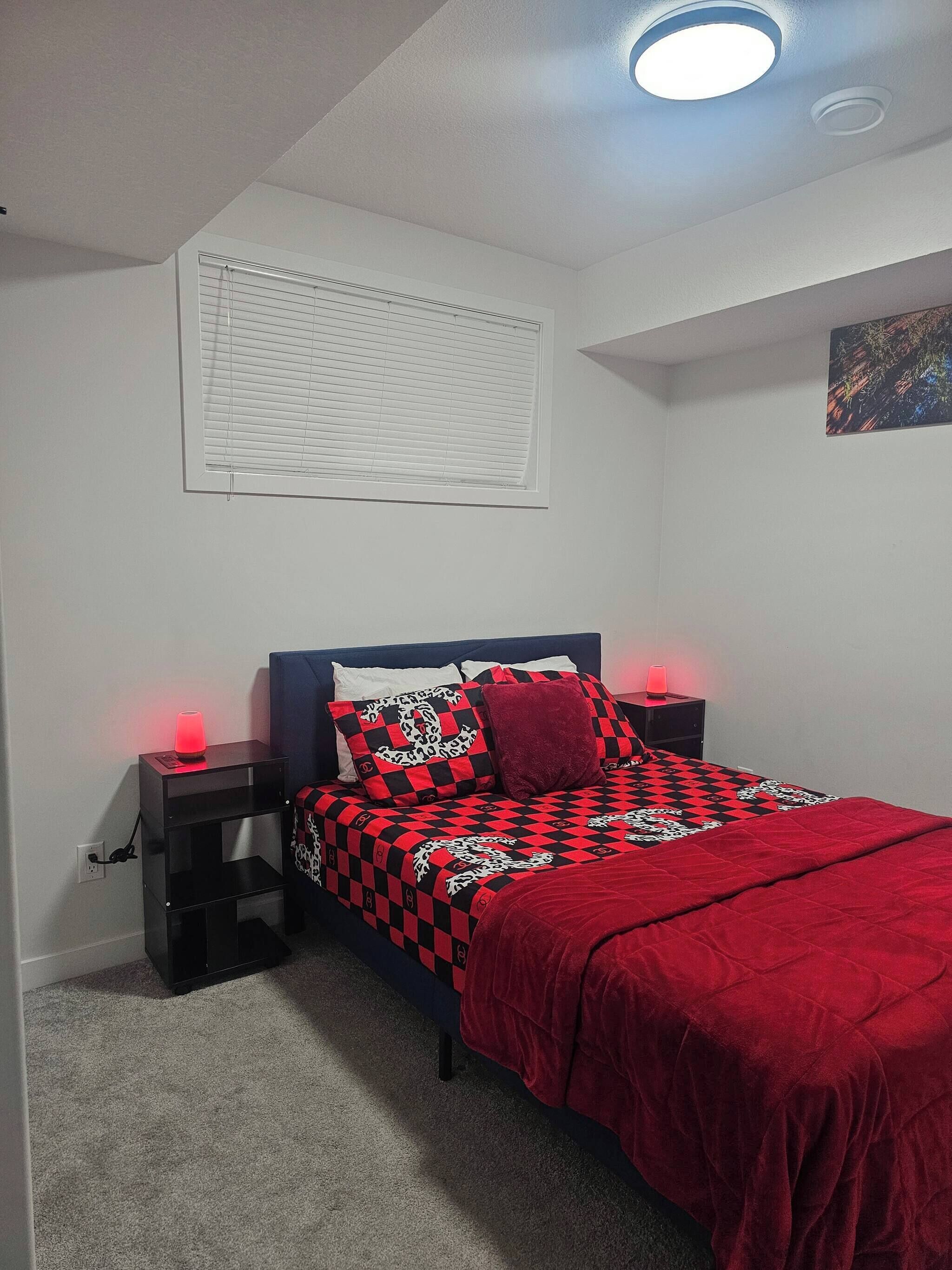 A bedroom with a bed, two bedside tables, a ceiling light, a window with blinds, and a wall-mounted artwork.